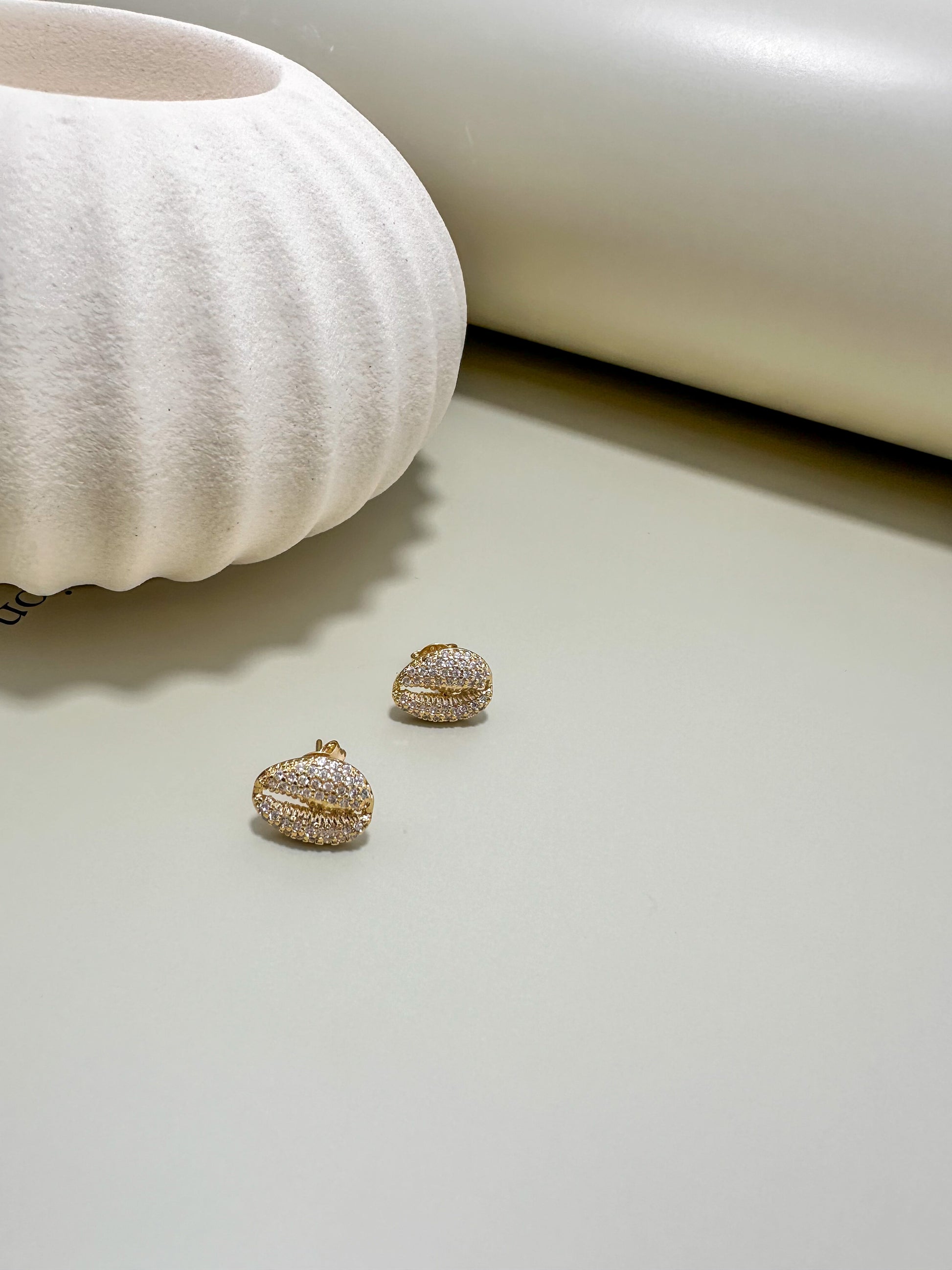 Velani Jewelry Cowrie Stud Earrings displayed beside a decorative white vase on a sleek surface, showcasing their sparkle.