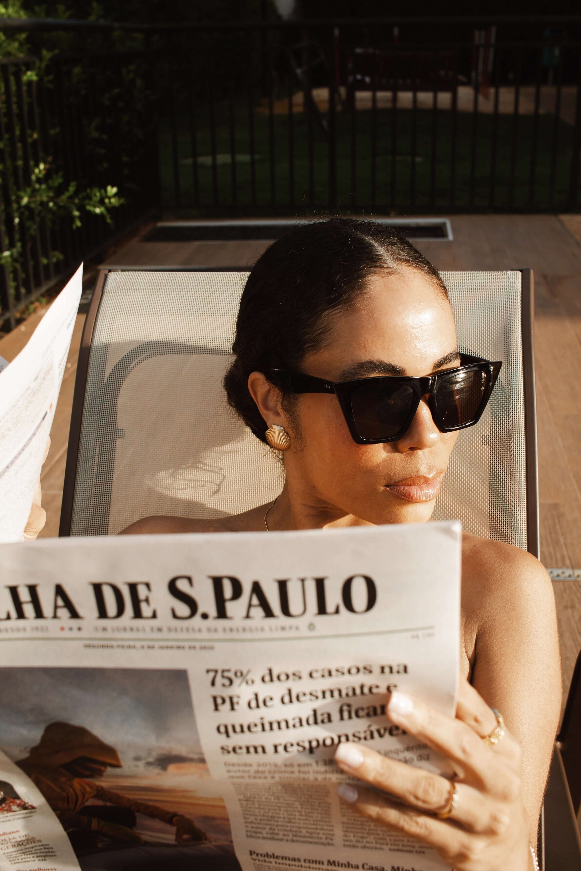 Woman wearing sunglasses reading a newspaper, adorned with Velani Jewelry clam shell stud earrings by the pool.