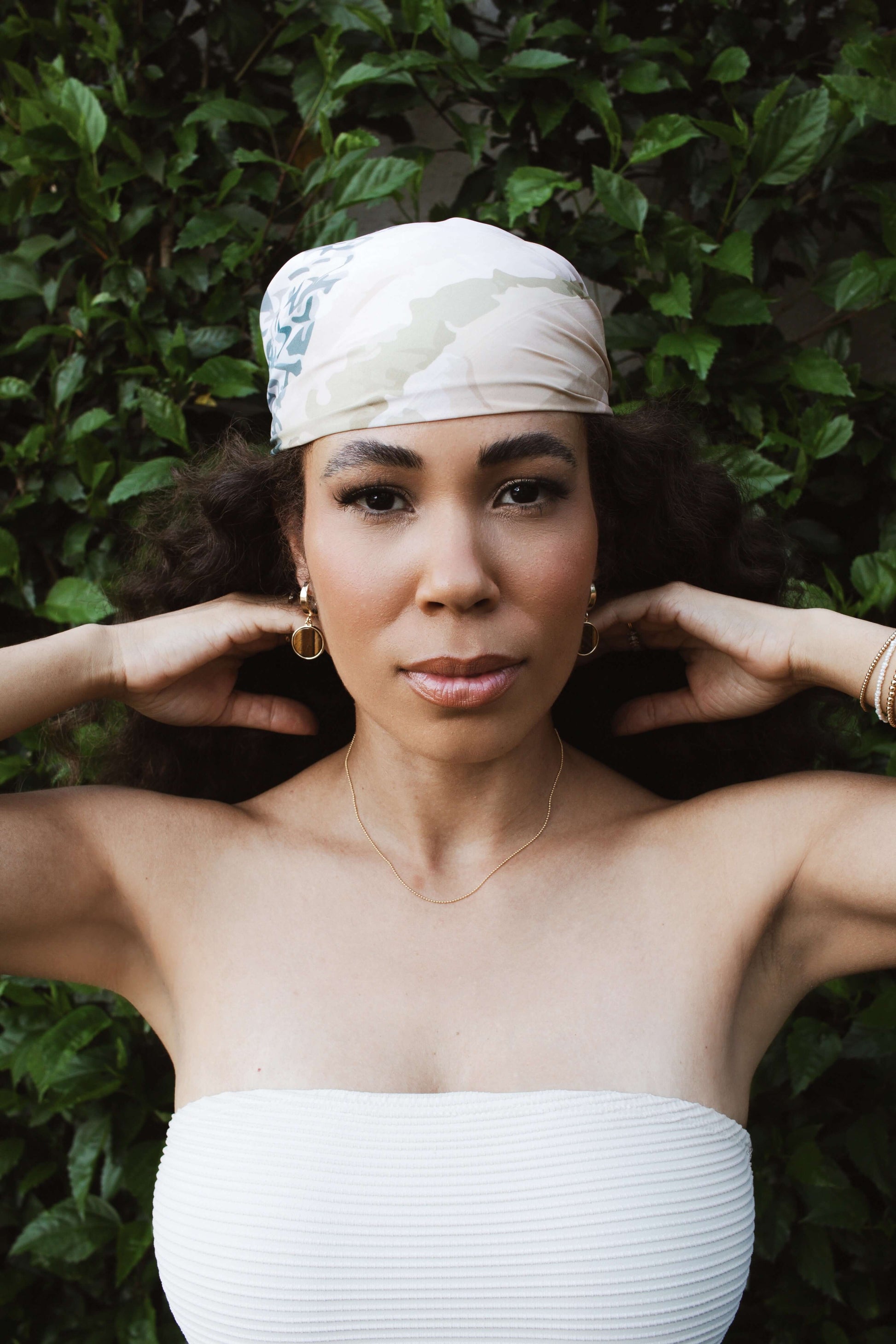 Woman with curly hair wearing a headscarf, posing gracefully against a backdrop of green foliage.